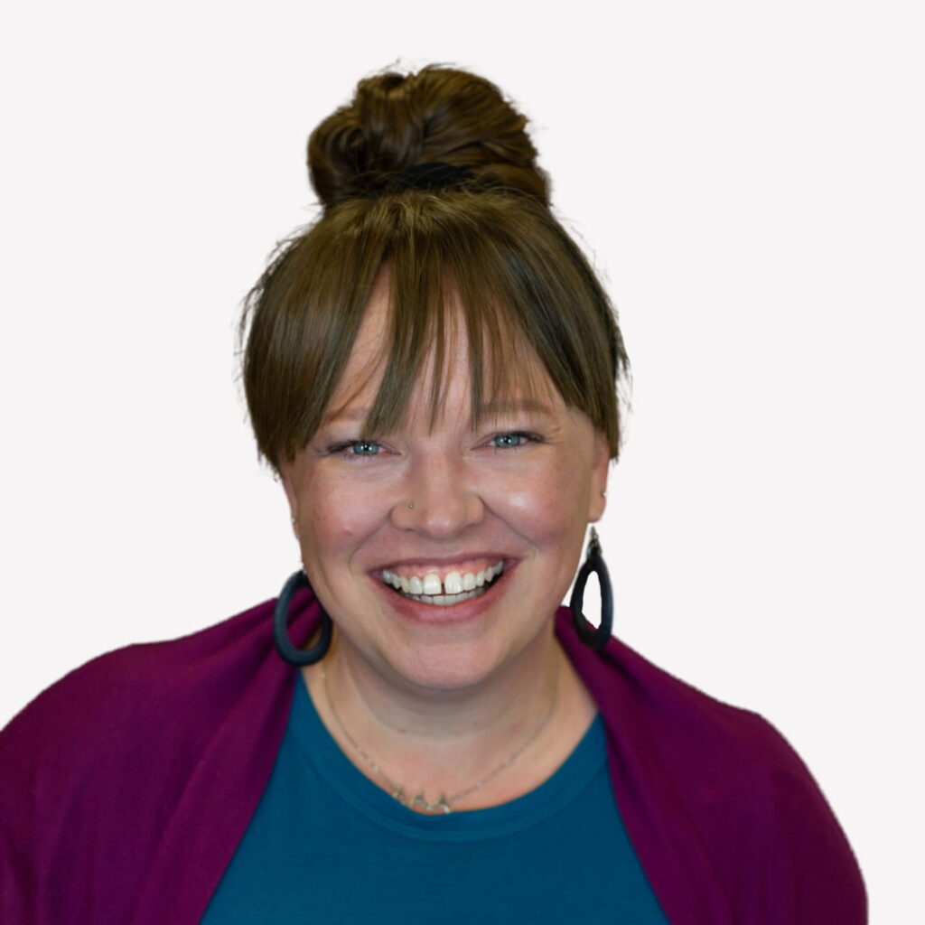 Woman with light skin smiling brightly, wearing a teal shirt and magenta cardigan, dark hoop earrings, and a necklace, with her brown hair styled in a top bun and straight bangs.