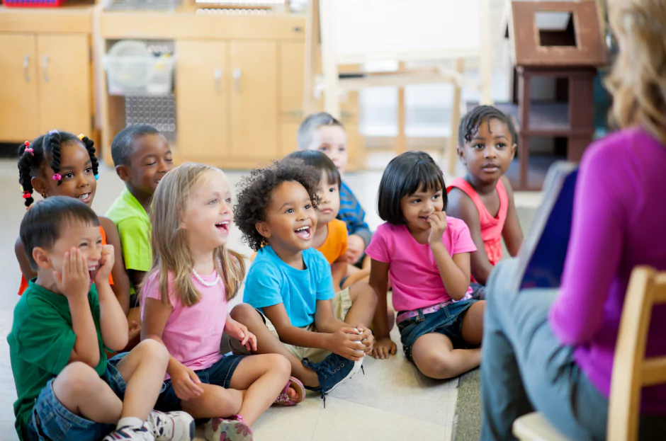 Diverse group of young children sitting on the floor smiling and listening to a teacher in a classroom setting during a children’s ministry lesson.