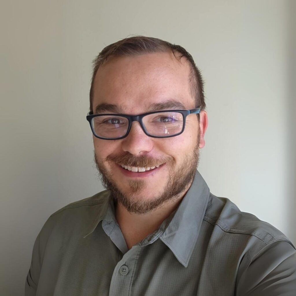 A man smiling in front of a plain light-colored wall, wearing glasses and a button-up shirt.