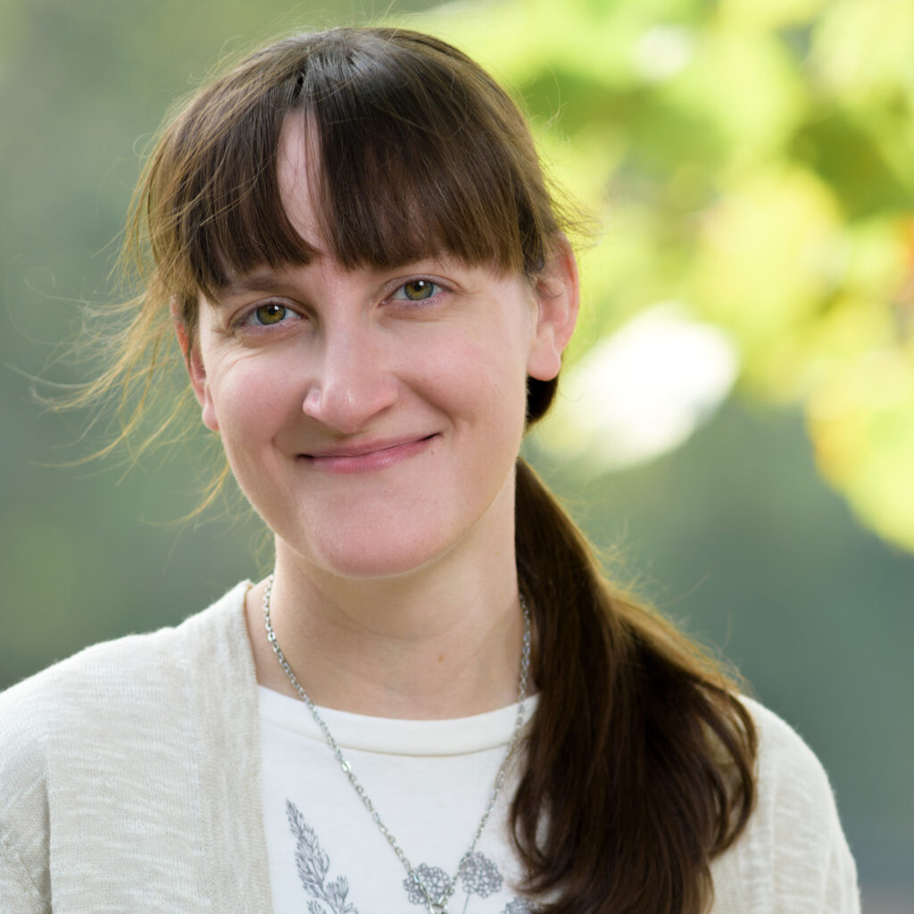 Woman smiling outdoors with sunlight filtering through green leaves in the background.