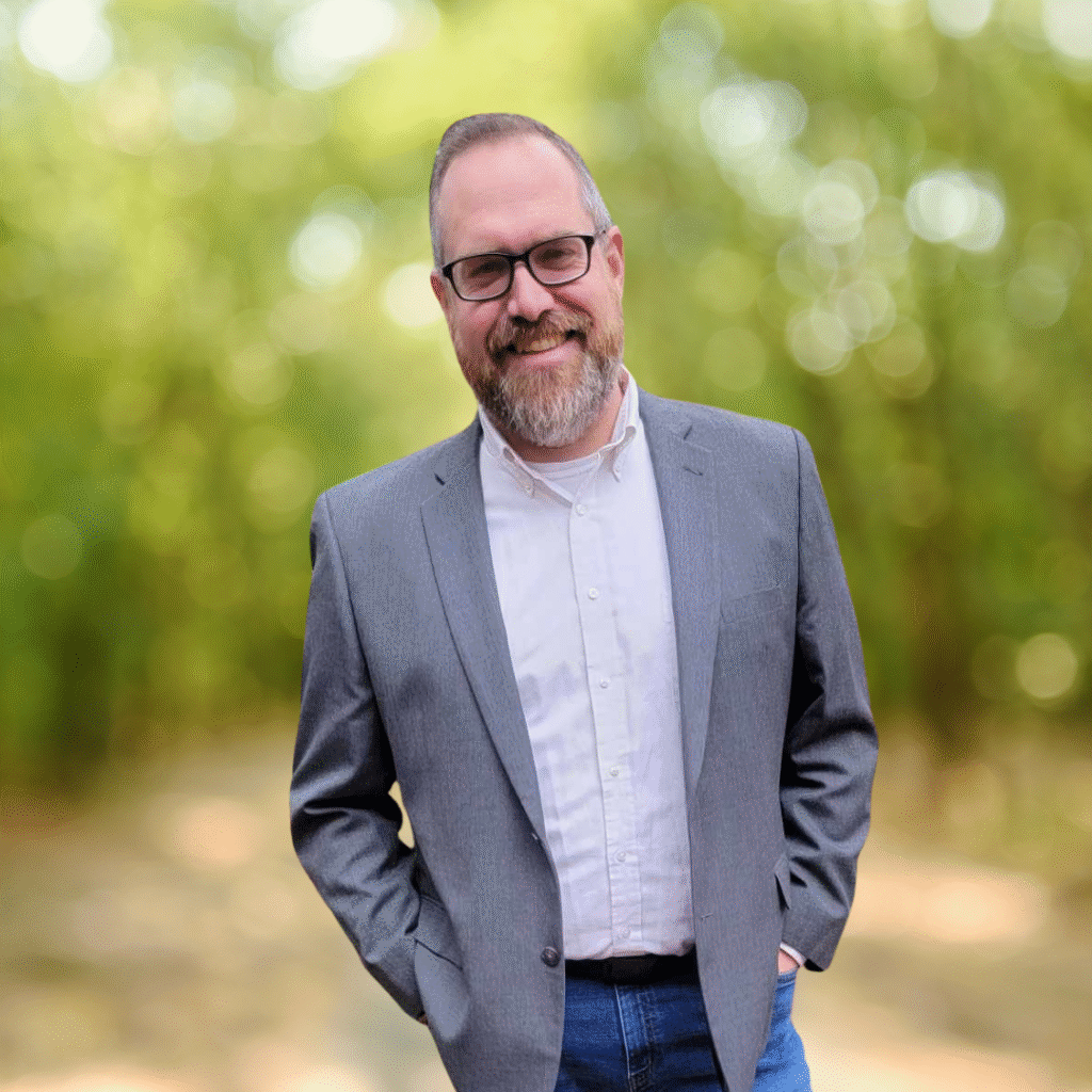 Man with a beard and glasses wearing a gray blazer and white shirt, standing outdoors with a soft, green, blurred background and smiling at the camera.