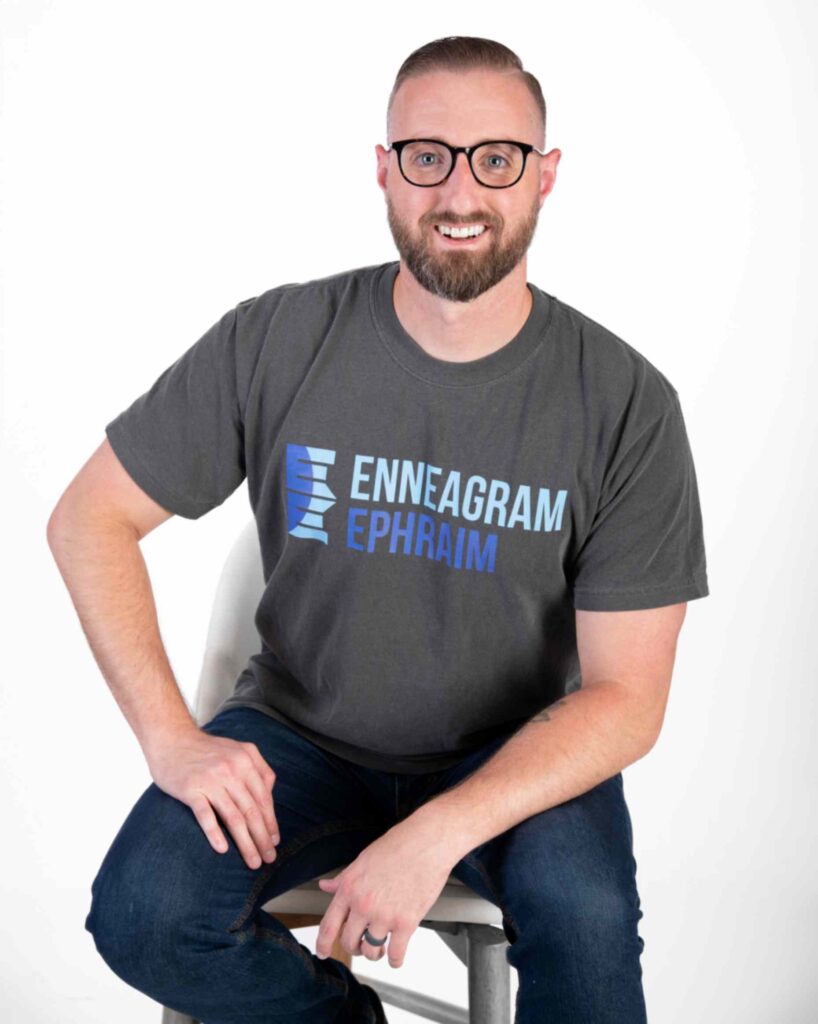 A smiling man with a beard and glasses sits on a white chair, wearing a dark grey t-shirt featuring the "Enneagram Ephraim" logo in white and blue text.