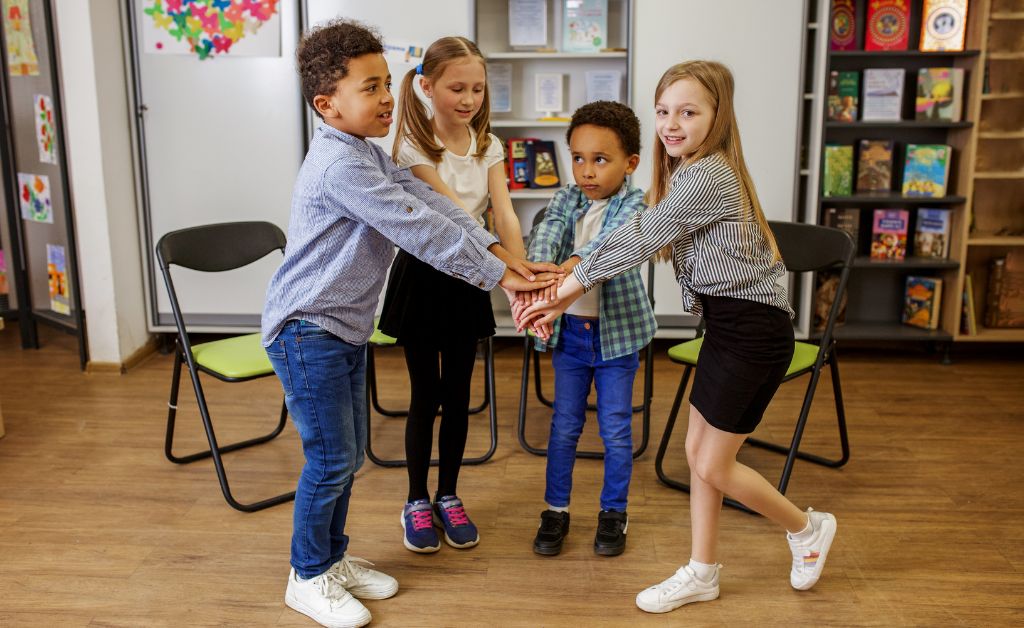 Four children stacking hands together in a circle during a low-key indoor youth group game in a classroom setting.